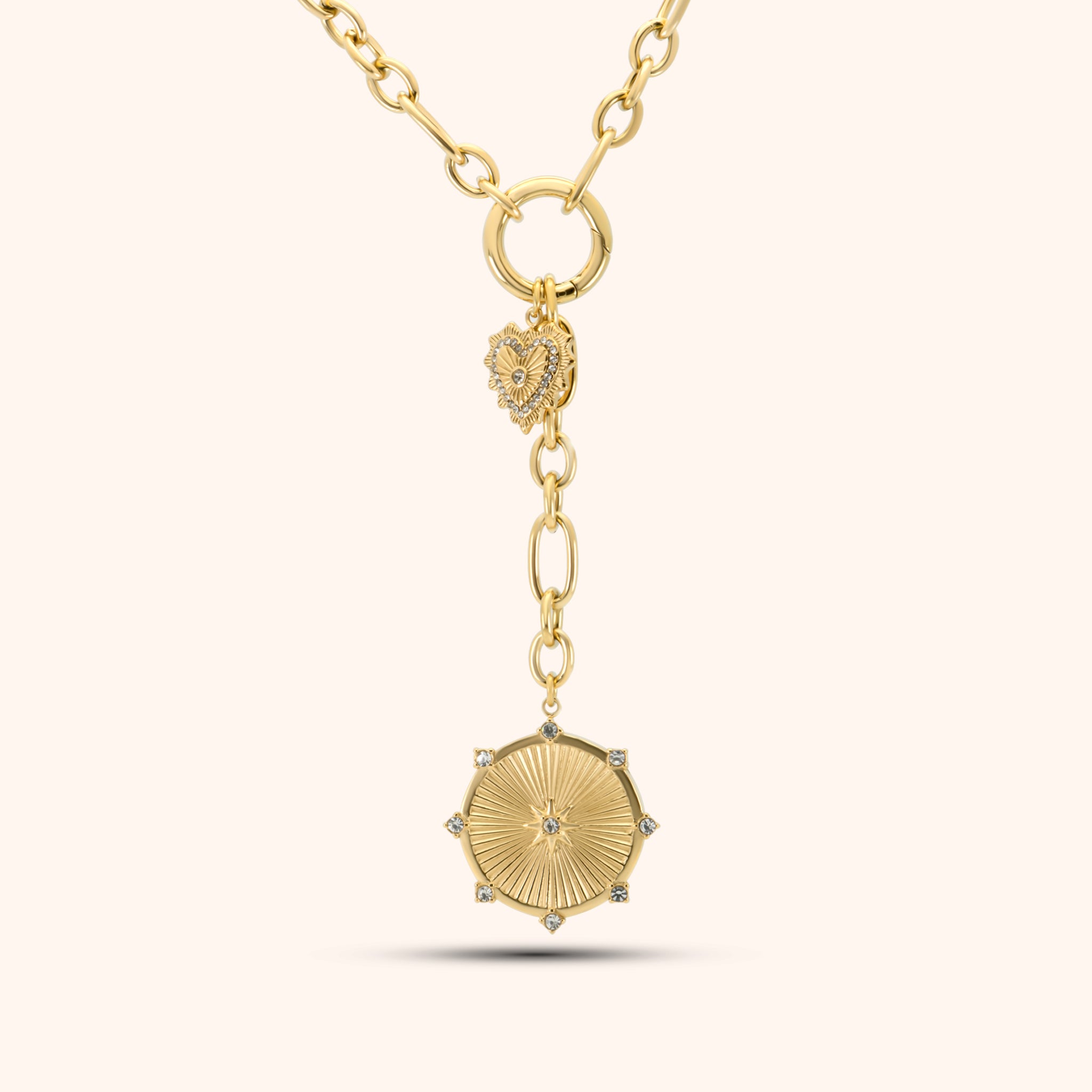 PRESSIE Radiant Heart Lariat necklace in 18k gold plate; features mixed paperclip chain, a central circular connector with a pavé heart charm, and a lariat drop ending in a sunburst medallion with clear stone accents.